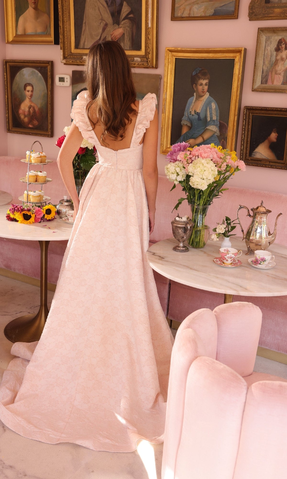 A woman in a long, light pink Ava Presley 40095 A-line prom dress stands with her back to the camera in a vintage-style room decorated with floral arrangements, tea sets, and portraits on pink walls.